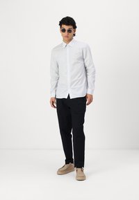 Camisa de rayas blanca y azul de manga larga, combinada con pantalones negros y zapatos beige. Modelo que lleva gafas de sol redondas.