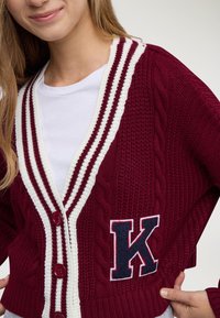 Giovane che indossa un cardigan lavorato a maglia color bordeaux con strisce bianche e una grande patch navy a forma di "K" sul davanti, sopra una t-shirt bianca.