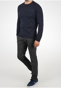 Maglione a maglia navy con collo tondo, finitura testurizzata, abbinato a pantaloni grigi scuri a gamba affusolata e sneakers in suede blu con suola bianca.