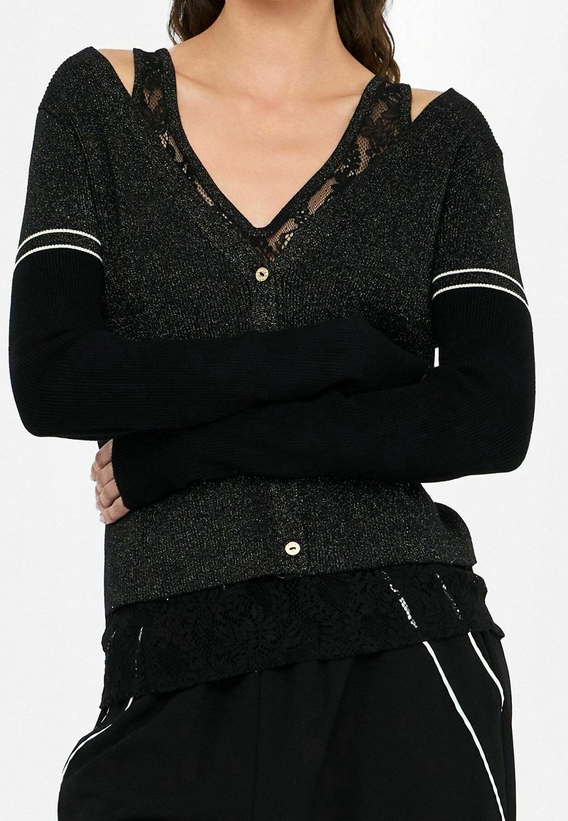 Femme portant un cardigan noir scintillant avec bordure en dentelle et boutons crème, associé à un pantalon noir avec des bandes blanches sur les côtés.