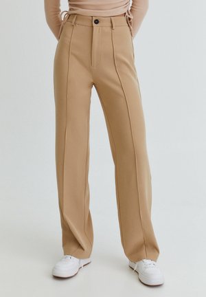 Beige bukser med høj talje og lige ben med forreste sømme, båret med hvide sneakers og en lysebeige langærmet top.