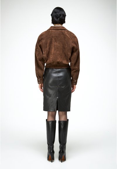 Personne aux cheveux courts et foncés, vue de dos, portant une veste en suède marron, une jupe crayon en cuir noir, des collants transparents et des bottes noires montant jusqu'aux genoux.