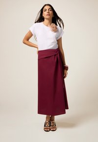 Camiseta blanca con detalle de flor de tela, combinada con una falda larga de wrap en color burdeos. Lleva sandalias negras con acentos metálicos.