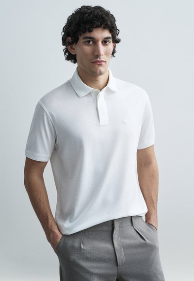 Polo blanco con cuello texturizado y un pequeño logo bordado; combinado con pantalones grises y entallados. Acabado de tela lisa.