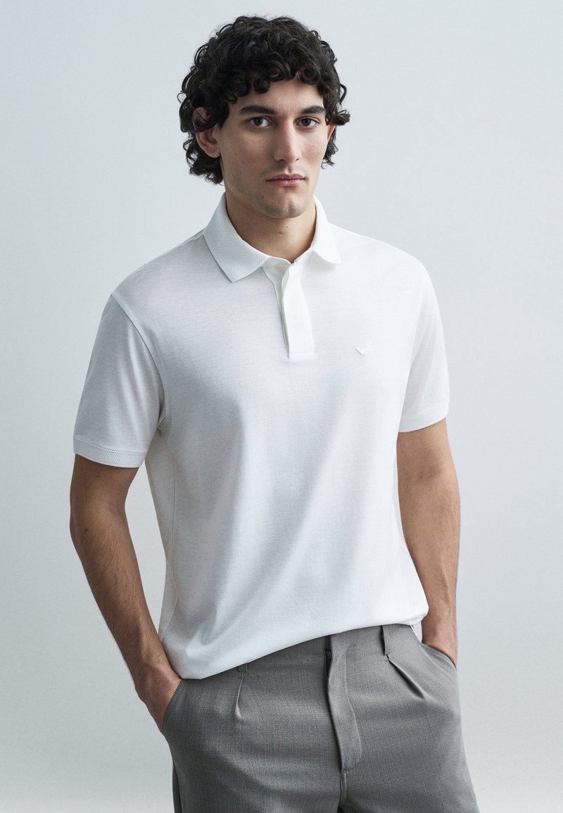 Bílá polokošile s texturovaným límečkem a malým vyšitým logem; doplněná o šedé, na míru šité kalhoty. Hladký povrch látky.