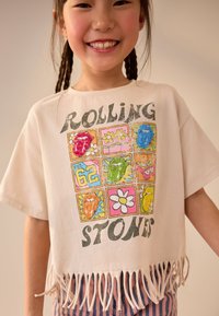 Fehér, rövidített póló rojtos szegéllyel. Többszínű grafikus négyzeteket ábrázol, amelyek ajkakat és virágokat mutatnak. Feltűnő felirat: "ROLLING STONES."