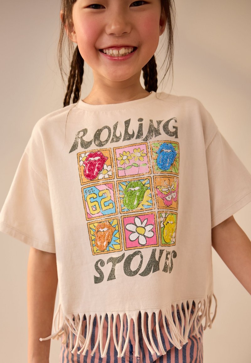 T-shirt blanc court avec ourlet à franges. Présente des carrés graphiques multicolores montrant des lèvres et des fleurs. Texte en gras : « ROLLING STONES ».