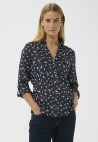 Femme blonde portant un blouse à manches longues bleu marine à imprimé floral blanc et un jean bleu foncé, les mains dans les poches, fond neutre.