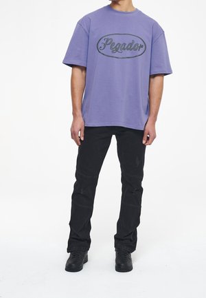 Hombre vestido con una camiseta morada holgada de "Pegador", jeans negros desgastados y zapatillas negras, de pie contra un fondo blanco liso.