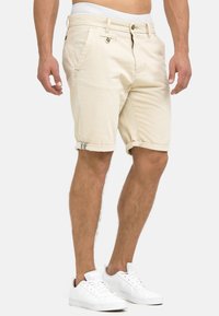 Lichtbeige katoenen shorts met opgerolde manchetten, voorzakken en een knoopsluiting. Gemaakt in combinatie met witte sneakers. Minimalistisch ontwerp.