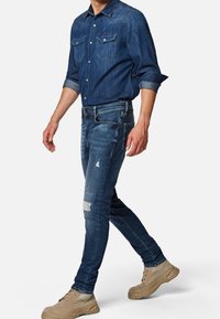 Men's denim shirt in dark blue with button-down front and dual chest pockets, paired with faded blue skinny jeans featuring distressed details.