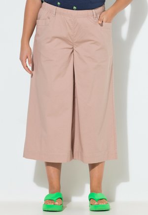 Osoba nosząca luźne, beżowe spodnie 3/4 z kieszeniami, granatowy top z małymi nadrukami palm i jaskrawozielone sandały typu slide.