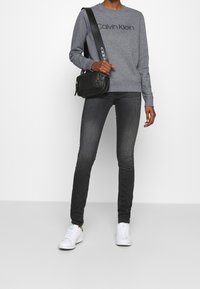 Sudadera gris con el logo negro de "Calvin Klein", combinada con jeans oscuros. Zapatillas blancas y un pequeño bolso cruzado negro. Atuendo simple y casual.