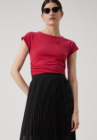 Red fitted t-shirt with short sleeves featuring a metallic logo at the chest, paired with a black pleated skirt with a branded waistband.