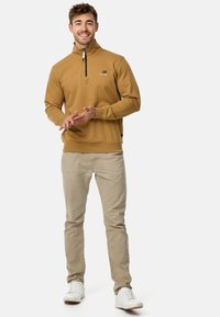 Sudadera de media cremallera en color mostaza, hecha de material suave, con un logo en el pecho y puños acanalados, combinada con pantalones beige claros.