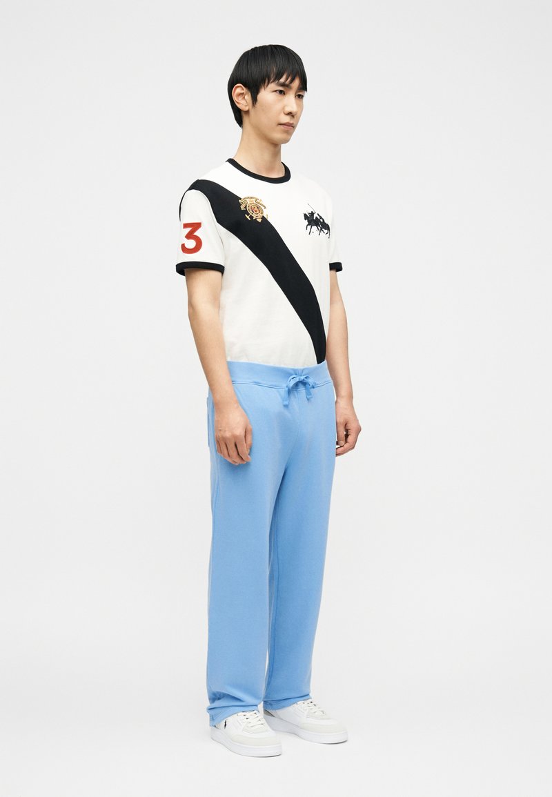 Jeune homme portant un polo blanc avec une bande diagonale noire, un numéro 3 rouge sur la manche, un pantalon de survêtement bleu clair et des baskets blanches, debout.