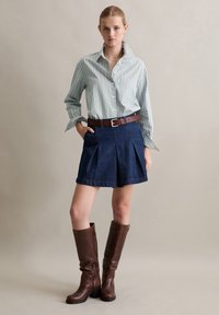 Camicia a maniche lunghe striata verde e bianca, gonna di jeans denim a pieghe blu scuro, cinta in pelle marrone e stivali al ginocchio in pelle marrone.