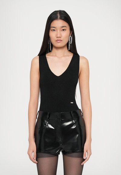 Femme aux longs cheveux noirs portant un haut noir sans manches en V, un short noir brillant, des collants noirs transparents et des boucles d'oreilles en cristal pendantes.