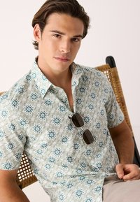 Camisa de manga corta en un patrón floral en verde claro y blanco, con acentos en azul y un cuello abotonado. Unas gafas de sol descansan sobre la camisa.