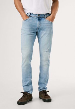 Mand iført lyseblå slim fit jeans med let slid, kombineret med mørkebrune casual sneakers, hænderne i lommerne.