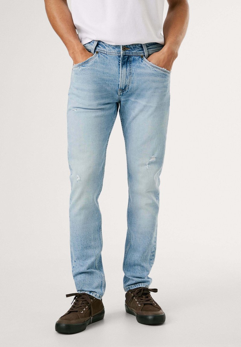 Homme portant un jean slim fit bleu clair légèrement usé, associé à des baskets décontractées marron foncé, les mains dans les poches.