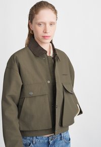 Olivengroen cropped jack met een ribbelkraag, drukknopen aan de voorkant en twee grote zakken. Gemaakt van duurzaam katoen.