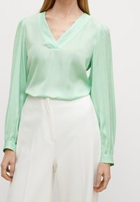 Blusa verde menta a maniche lunghe con scollatura a V e una texture morbida e liscia. Presenta maniche con polsini ed è abbinata a pantaloni bianchi a gamba larga.