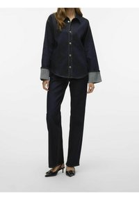 Mörk denim skjorta med knappar och förlängda manschetter, tillsammans med mörka raka jeans och svarta klackskor. Slät yta, klassisk design.