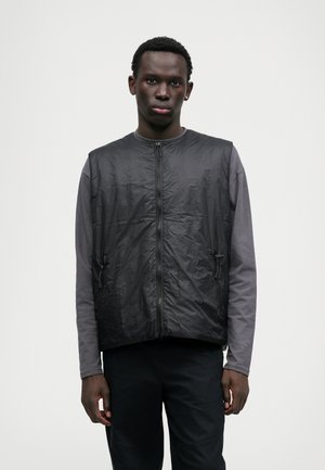 Gilet nero senza maniche con una superficie liscia e lucida, zip frontale e tasche laterali, indossato sopra una maglietta grigia a maniche lunghe.