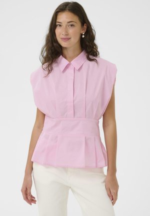 Žena s valovitom smeđom kosom koja nosi naušnice od bisera, bezrukavu svijetloružičastu bluzu s ovratnikom i kremaste hlače, stoji na bijeloj pozadini.