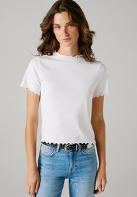 Λευκό ριμπ μπλουζάκι με κυματιστή λαιμόκοψη και άκρες μανικιών, σε συνδυασμό με μπλε τζιν και μαύρη ζώνη με τρουκς.