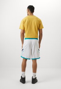 Yellow short-sleeve shirt; white sports shorts with teal accents and pockets. Black athletic shoes and white socks. Plain background.