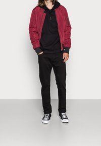 Giacca bomber rossa con polsini a costine sopra un hoodie nero, abbinata a pantaloni neri e sneakers bianche e nere. Materiale liscio, vestibilità comoda.