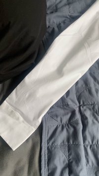 Camicia a maniche lunghe realizzata in tessuto bianco, texture liscia, con polsini puliti. Tessuto nero parzialmente visibile nell'angolo in alto a sinistra.