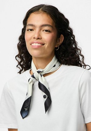 Écharpe en soie crème avec des pois noirs, nouée autour du cou. Portée sur un t-shirt blanc uni, présentant une texture lisse et une finition brillante.