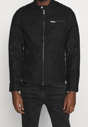 Mand iført en sort jakke med lynlås og sorte jeans med hænderne afslappet langs siderne mod en ensfarvet baggrund.