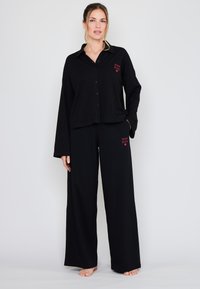 Musta loungevaate-setti, johon kuuluu napillinen paita, jossa on kaulus, ja leveälahkeiset housut. Molemmissa osissa on huomattavat punaiset logopainatukset. Pehmeä kangas.