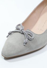 Chaussure plate en suède gris avec des pois blancs, bout pointu et un nœud décoratif gris à l'avant. Intérieur lisse et extérieur texturé.
