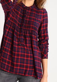 Chemise à carreaux rouge et marine, avec manches longues, détails plissés sur le devant et coupe décontractée. Tissu en coton à la texture douce.