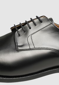Chaussure en cuir noir avec finition lisse, dotée de cinq œillets pour les lacets, de coutures contrastées et d'un bout arrondi.