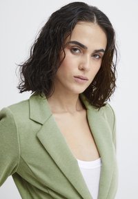 Light green blazer with a notched collar, textured fabric. Layered over a white top, featuring a fitted silhouette and structured shoulders.