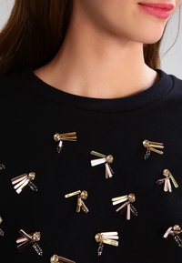 Felpa nera con decorazioni metalliche che sembrano forcine e perline, creando un motivo testurizzato attraverso il tessuto.