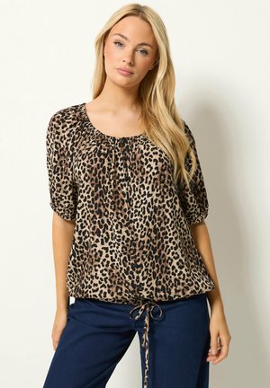 Blond vrouw met lang haar die een luipaardprint blouse met pofmouwen en donkerblauwe broek draagt, staande tegen een effen achtergrond.