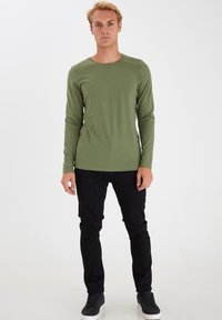 Langærmet olivengrøn skjorte i blødt, strækbart materiale parret med sorte slim-fit jeans og sorte sneakers med hvide såler.
