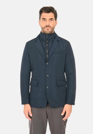 Uomo con capelli scuri e barba che indossa un blazer blu navy con bottoni sopra una giacca con zip e collo alto e pantaloni grigi, in piedi davanti a uno sfondo bianco.