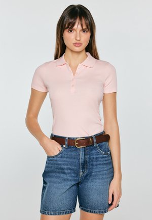 Kobieta ubrana w jasnoróżową koszulkę polo z krótkim rękawem wpuszczoną w wysokodźwiękowe niebieskie szorty jeansowe z brązowym pasek.