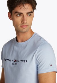 Light blue cotton t-shirt featuring "TOMMY HILFIGER" logo in dark navy, established year 1985, with contrasting logo accent on sleeve.