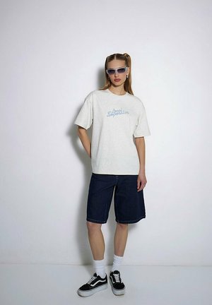 Jasnoszara koszulka z niebieskim napisem, szorty z ciemnego dżinsu do kolan, białe skarpetki i czarne sneakersy. Model nosi okulary przeciwsłoneczne.