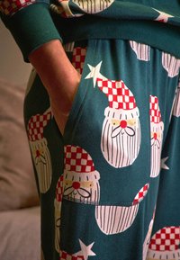 Zelené pyžamové nohavice s bielymi, červeno-kockovanými tvárami Santa Clausa s výraznými bradami a červenými nosmi, vyrobené z mäkkej, elastickej látky.