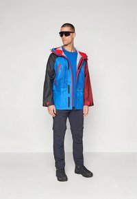 Blå och röd vattenavvisande jacka med svarta ärmar, flera fickor med dragkedja, kombinerad med marinblå cargobyxor och svarta vandringskängor.